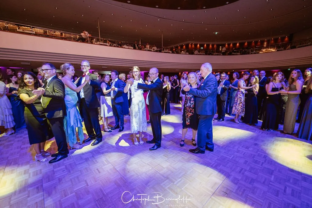 Viele Menschen tanzen in einem festlich dekorierten Saal, vermutlich bei einer Hochzeit oder Feier, mit Lichtern und einem Mehrstöckigen Balkon im Hintergrund.