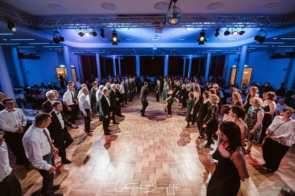 Viele Menschen tanzen in einem festlich geschmückten Saal mit Holzparkett, blauen Lichtakzenten und einer Tanzfläche in der Mitte bei einer Feier oder einem Event.