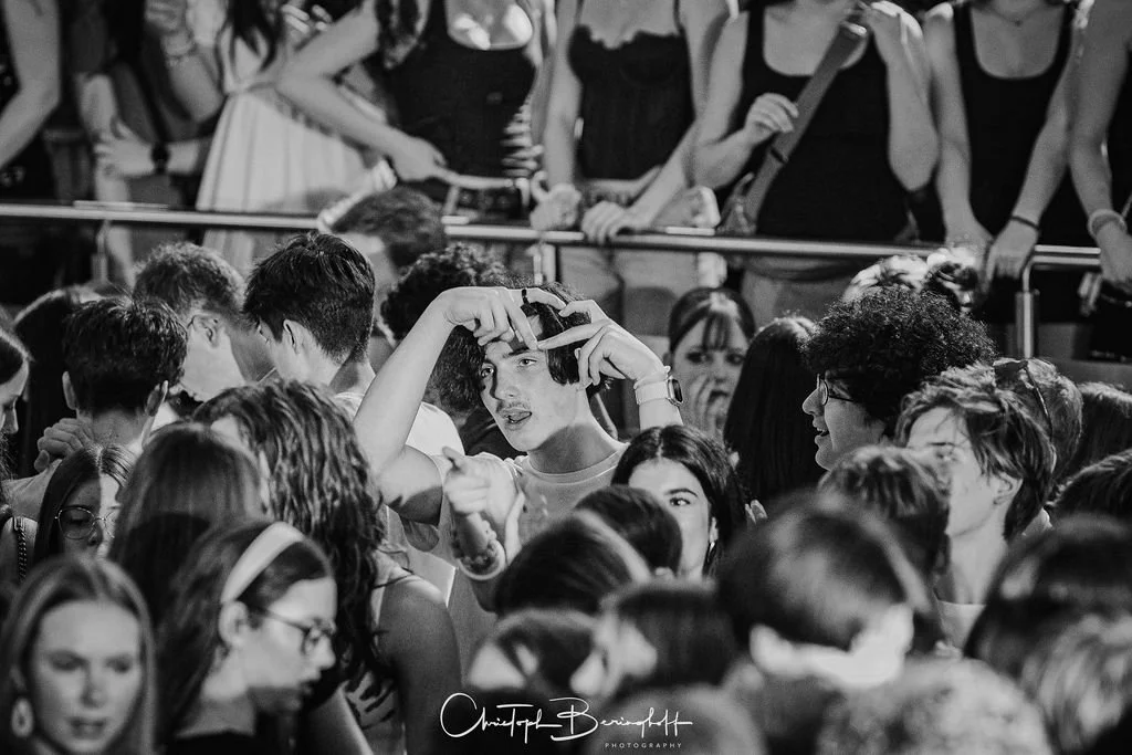 A crowd at an event; some people are taking photos; men and women of various ages; in the foreground, a boy taking photos with his cell phone; black-and-white photo