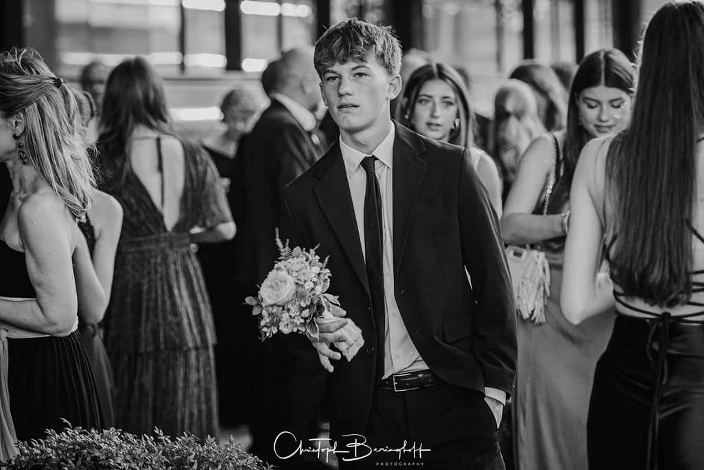 Junger Mann in formeller Kleidung mit Blumen bei einer Veranstaltung, umgeben von jungen Frauen und älteren Personen, im Innenraum.