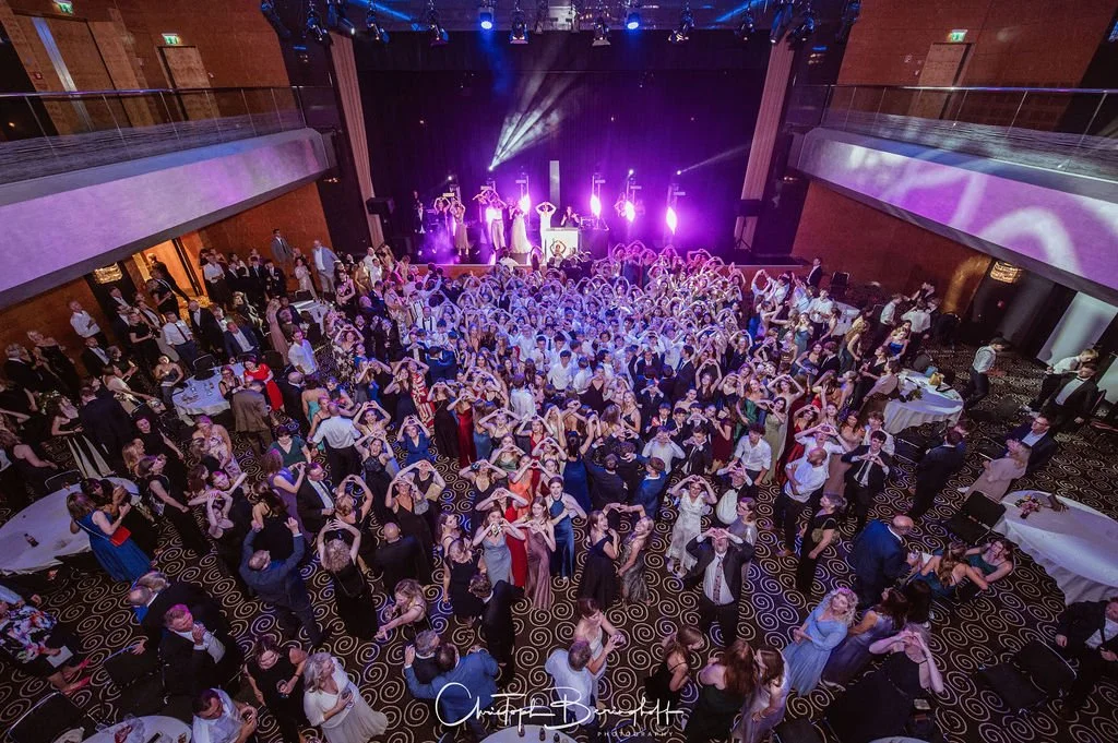 Große Tanzparty mit vielen Menschen auf der Tanzfläche, bunte Lichter und Musikbühne im Hintergrund, festliche Atmosphäre in einer großen Halle.