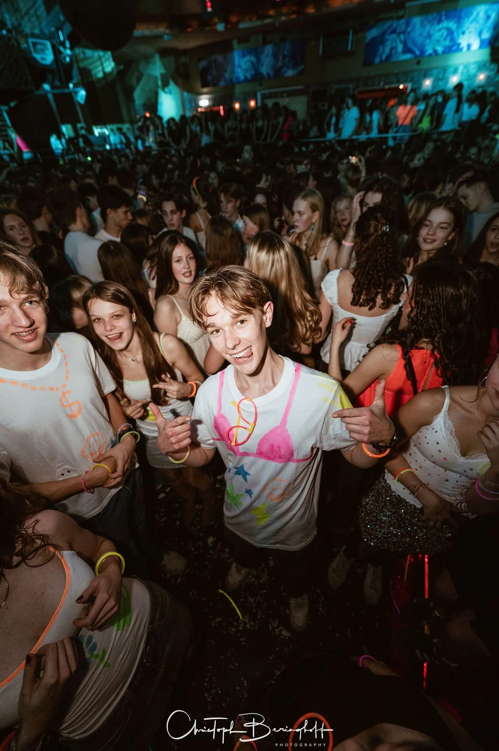 Young people are partying in a dark, noisy party hall with colorful lights and a large crowd in the background.