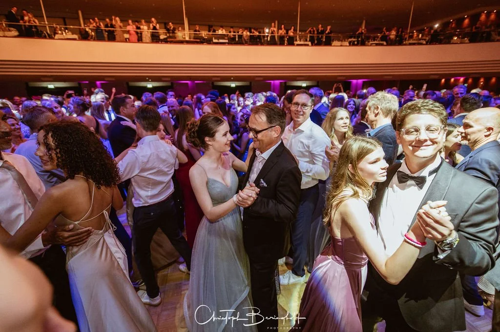 Menschen tanzen und feiern bei einer Veranstaltung, vermutlich ein Ball oder eine Party, in einem großen Saal mit Tanzfläche und Bühne, mit bunten Lichtern und einem Balkon mit Zuschauern