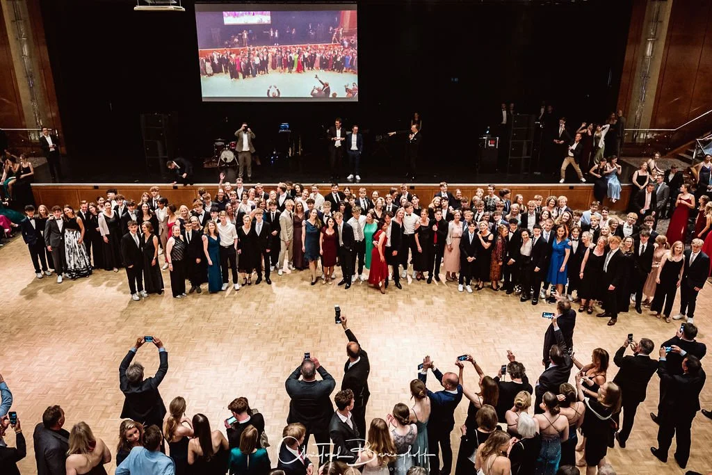 Gruppenfoto bei einer formellen Veranstaltung in einem großen Saal, viele Menschen in Abendkleidung, einige auf der Bühne, andere im Publikum, die Fotos machen.