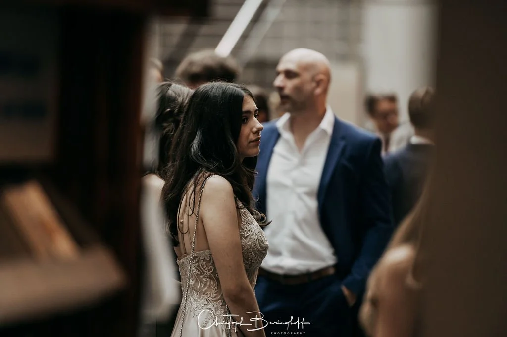 Eine junge Frau in einem eleganten Kleid steht in einem Raum voller Menschen, die im Hintergrund sprechen und sich unterhalten.