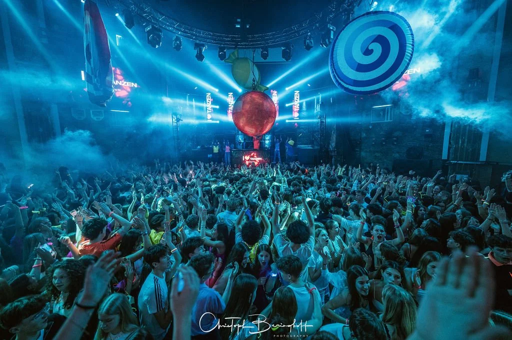 A large crowd at a party in a dark room with blue lighting; hanging in the center are large colorful decorations, such as a red light, a green ornament, and a large blue spiral candy. The people have their hands in the air and are celebrating.