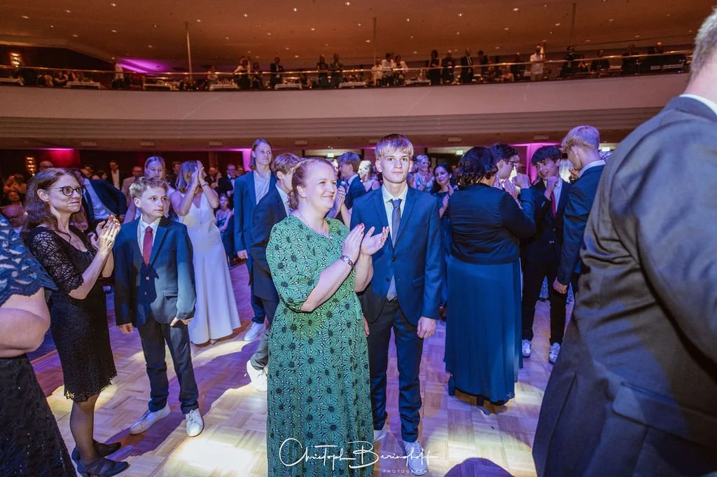 Menschen bei einer Tanzveranstaltung, die klatschen, im festlichen Anzug und Kleid auf der Tanzfläche in einem großen Saal mit mehreren Stockwerken.