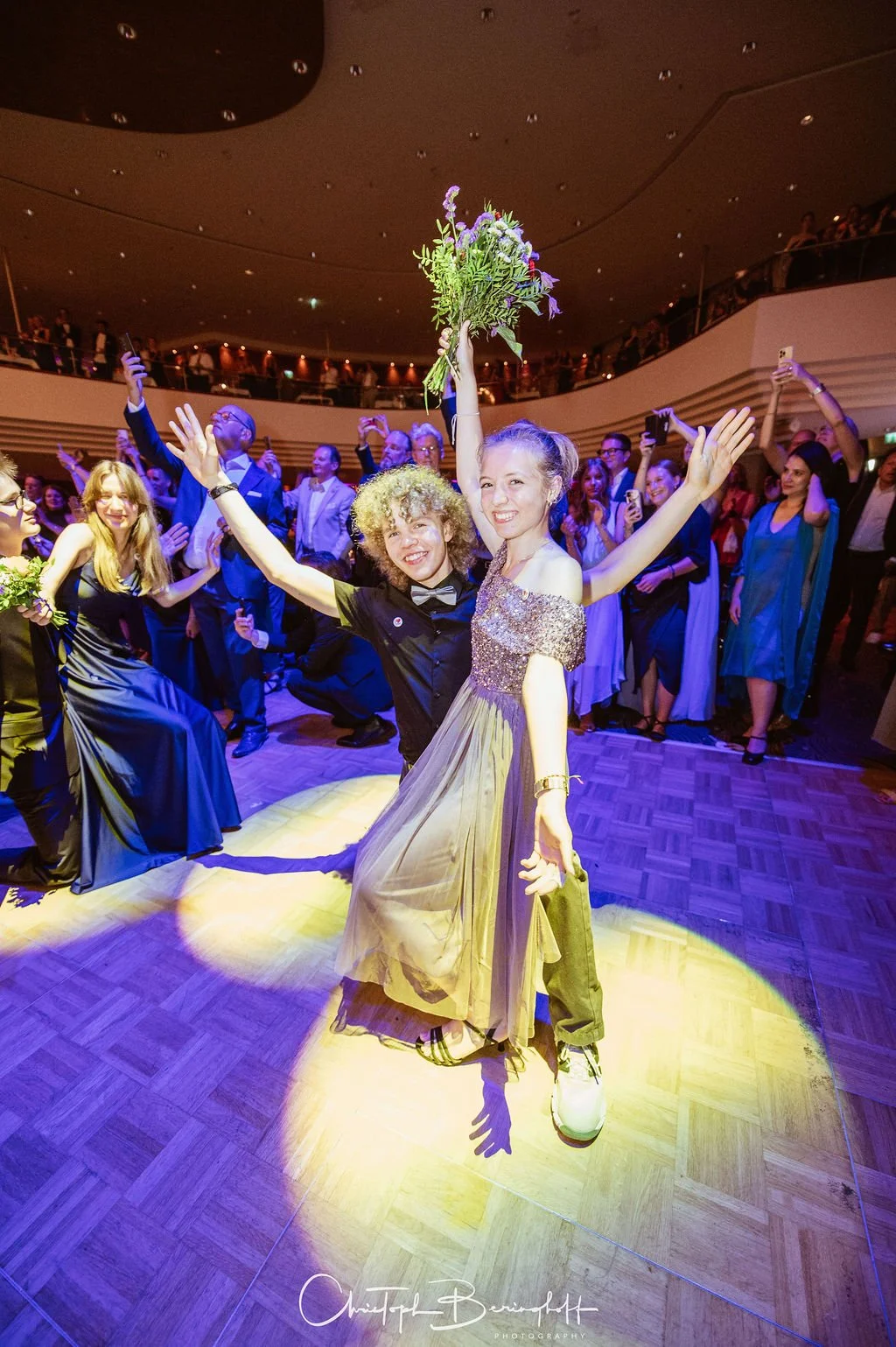 Freudengesteuerte Feier mit mehreren Menschen auf der Tanzfläche, zwei Frauen im Vordergrund, eine hält einen Blumenstrauß hoch, alle lachen und haben Spaß, im Hintergrund eine Menschenmenge und eine Treppe.
