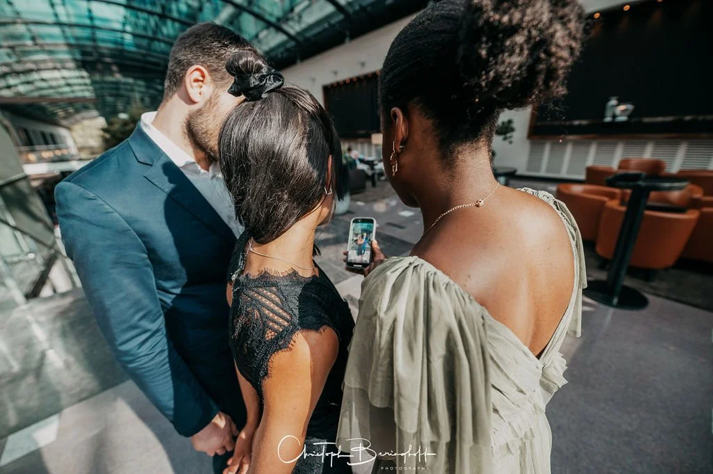 Drei Menschen schauen auf ein Smartphone, die Bilder im öffentlichen Innenraum machen, mit braunen Sitzgelegenheiten im Hintergrund.