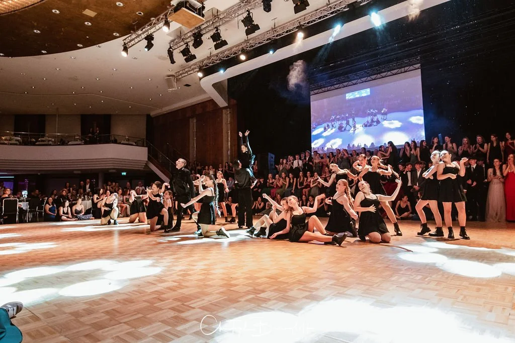 Tanzgruppe auf einer Bühne während einer Performance vor einem Publikum, mit einem großen Bildschirm im Hintergrund, in einem Veranstaltungsraum.