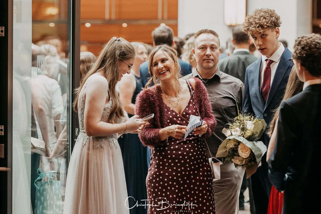 Menschen bei einer Feier, einige halten Blumen und Karten. Eine Frau in einem rot gepunkteten Kleid lacht, während eine andere junge Frau auf ihr Handy schaut.