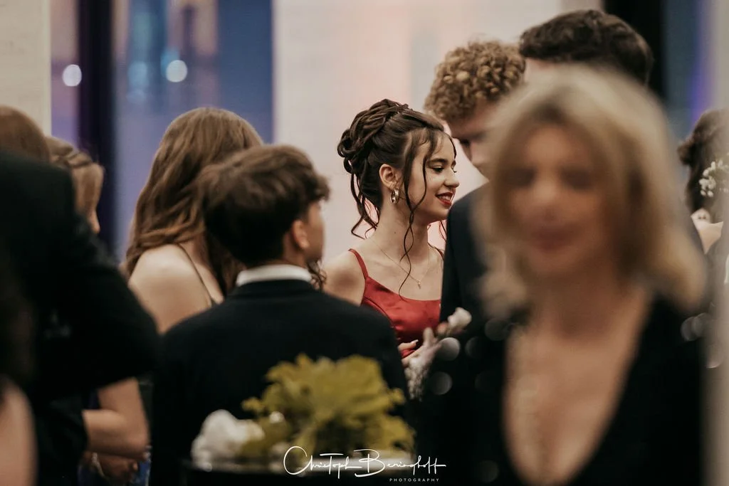 Menschen in formeller Kleidung bei einer Feier, junge Frau in rotem Kleid im Mittelpunkt, lachend, im Hintergrund verschwommen.