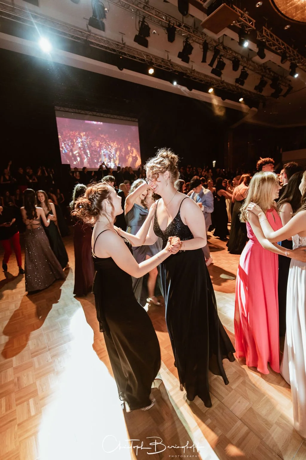 Menschen tanzen auf einem festlichen Ball, einige in eleganten langen Kleidern, in einem mit Scheinwerfern beleuchteten Saal.