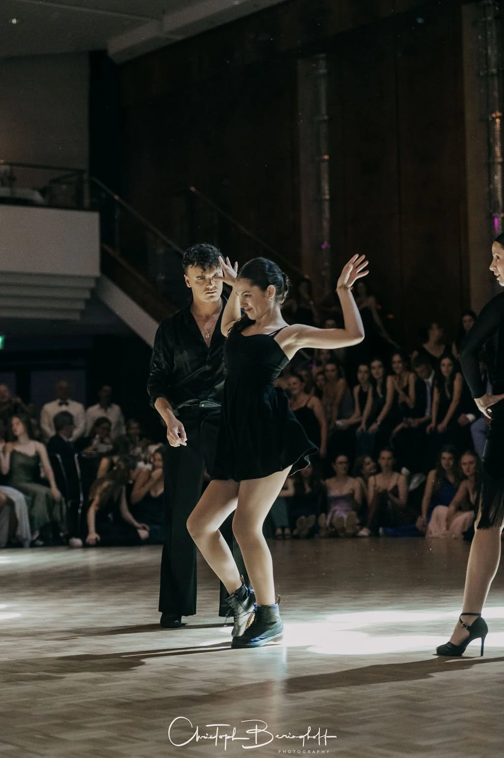 Paare tanzen in einem dunklen Raum vor einem Publikum, beim Tanzen im Stil des Tango, mit Fokus auf das Mädchen in der Mitte, das in einem schwarzen Kleid tanzt.