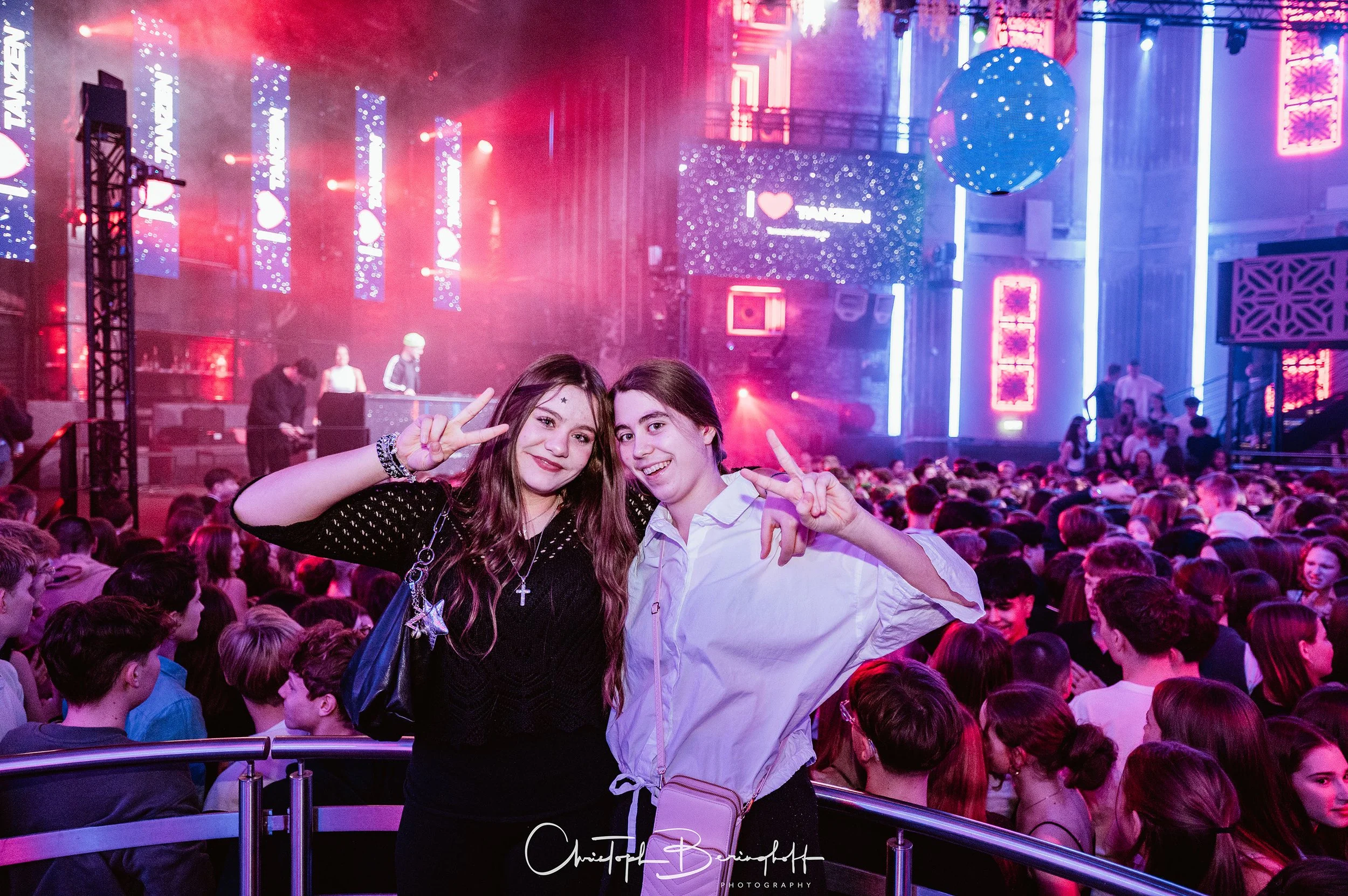 Zwei junge Frauen posieren bei einem Konzert oder Festival in einer großen, lebhaften Menge. Im Hintergrund ist eine Bühne mit bunten Lichtern und einer großen Leinwand zu sehen.