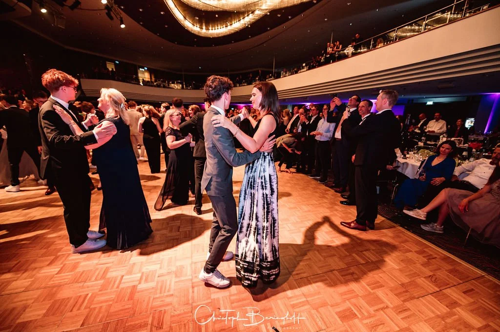 Tanzende Paare und Gäste auf einer Ballveranstaltung in einem festlichen, eleganten Saal mit Holzparkett und stimmungsvoller Beleuchtung.