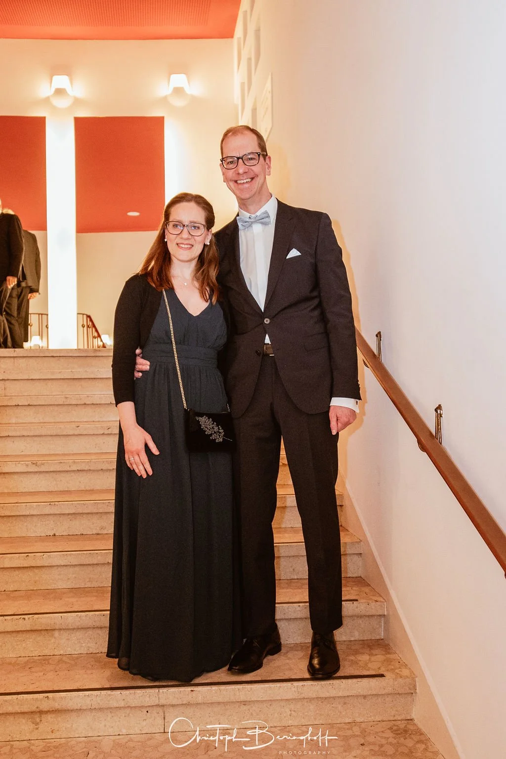 Ein Mann und eine Frau in formeller Kleidung, die zusammen auf einer Treppe stehen, bei einer Feierlichkeit im Innenraum.