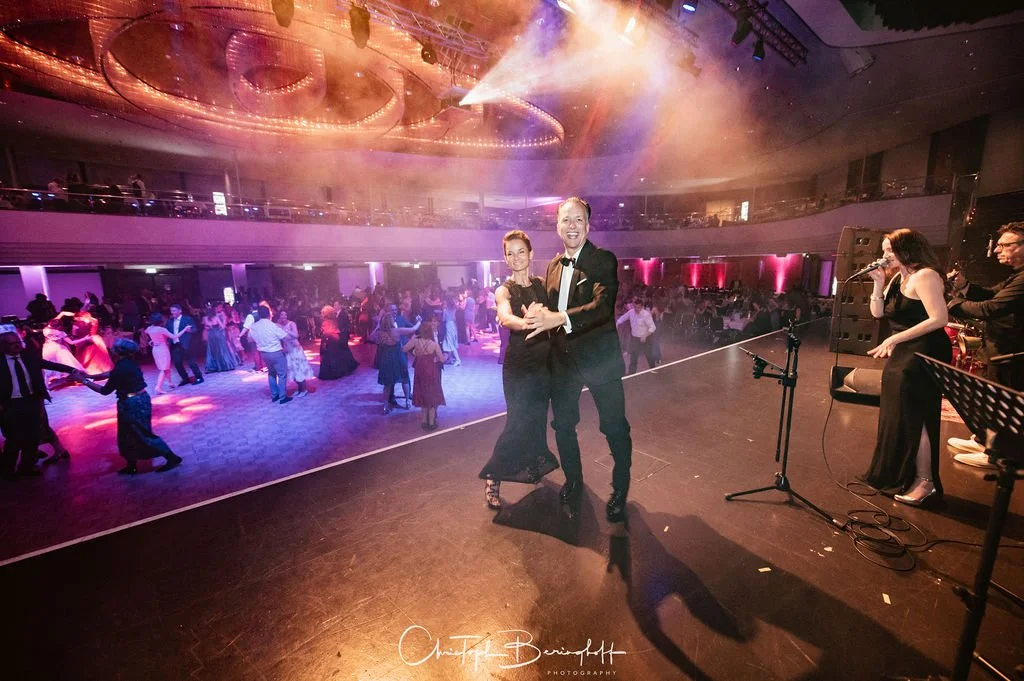 Tanzpaar auf der Bühne bei einer Ballveranstaltung, im Hintergrund tanzende Gäste in eleganter Kleidung, Musiker spielen live, bunte Lichteffekte.