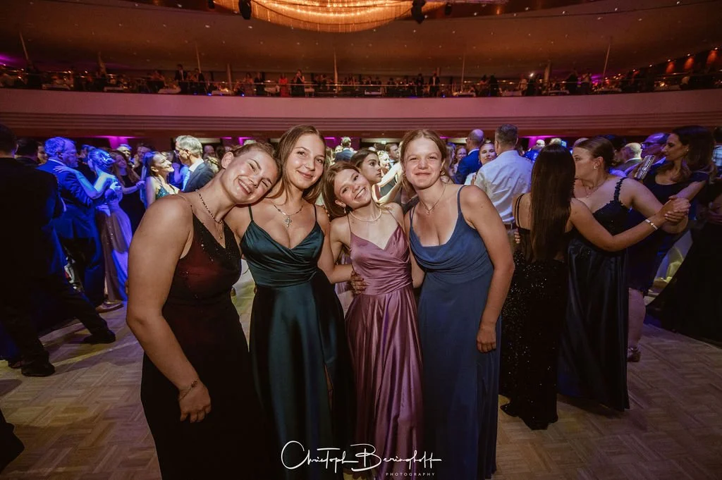 Vier junge Frauen in Abendkleidern auf einer Tanzfläche bei einer formellen Veranstaltung, umgeben von weiteren tanzenden Paaren. Hintergrund mit Lichteffekten und mehreren Ebenen des Veranstaltungsortes.