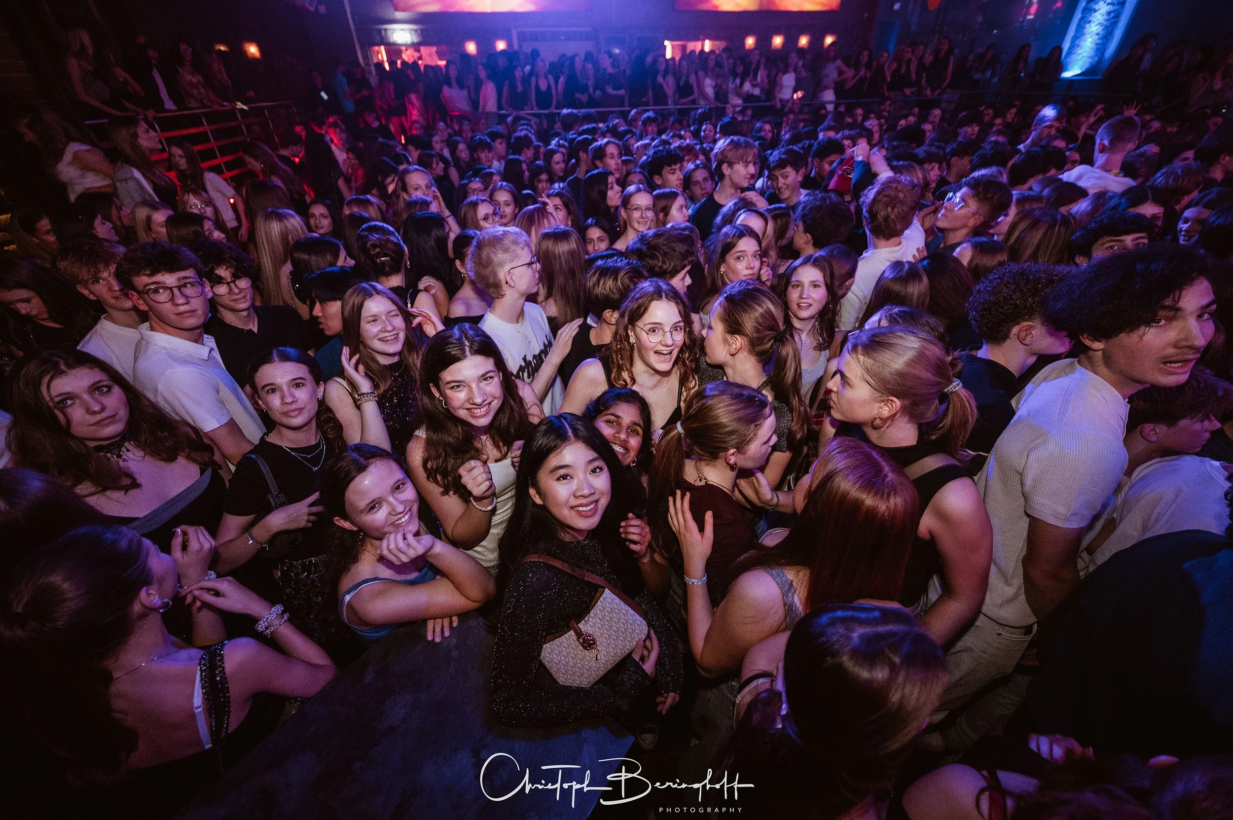 Vielzahl von Menschen in einem dunklen Raum bei einer Veranstaltung, einige lächeln in die Kamera, alle scheinen Spaß zu haben.