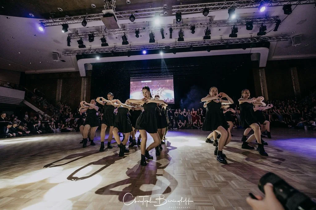 Tanzegruppe performing auf einer Bühne bei einer Veranstaltung, umgeben von Publikum, mit Rückenprojektor im Hintergrund.