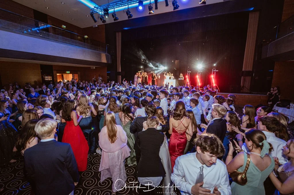 Junge Menschen in formeller Kleidung tanzen und feiern auf einer Tanzfläche bei einer Veranstaltung, mit einem DJ oder Band auf der Bühne im Hintergrund und bunter Bühnenbeleuchtung.