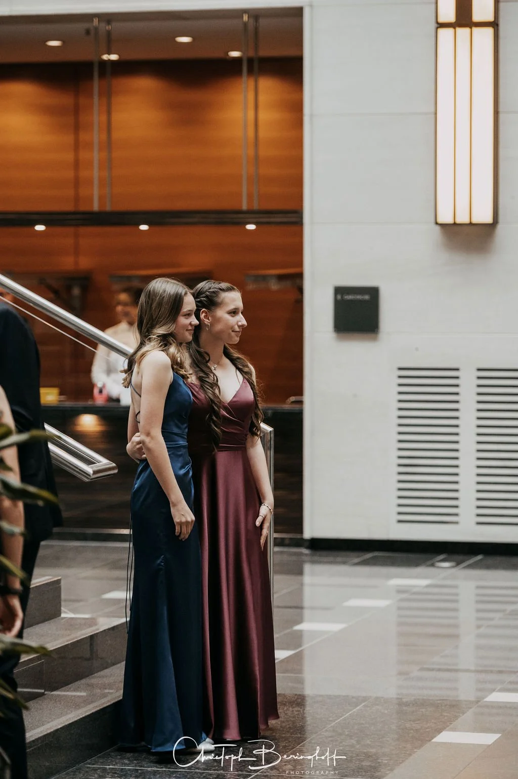 Zwei Frauen in elegante Abendkleider stehen nebeneinander im Inneren eines modernen Gebäudes, wahrscheinlich bei einer formellen Veranstaltung.