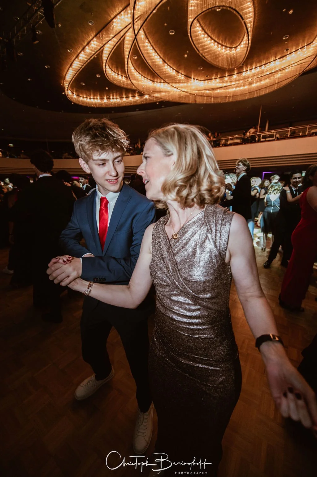 Ein junger Mann in blauer Anzug und roter Krawatte tanzt mit einer älteren Frau in einem glitzernden Kleid auf einer Party in einem großen Saal mit elegantem Deckenschmuck.