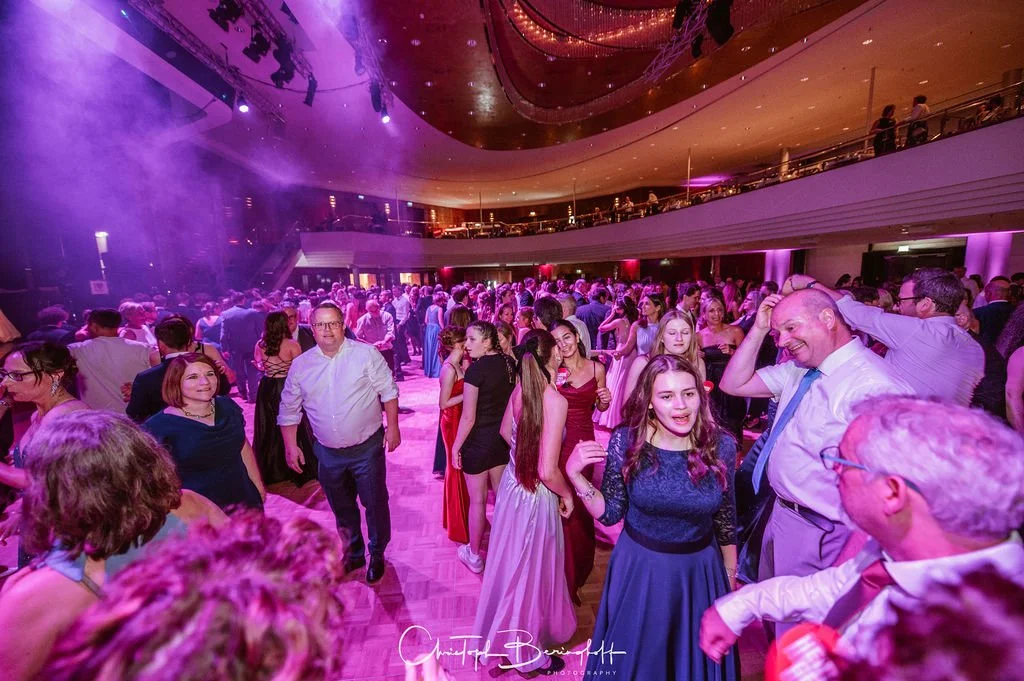 Menschen tanzen und feiern in einem festlich beleuchteten Saal, mit bunten Lichtern und einer zweiten Etage im Hintergrund.