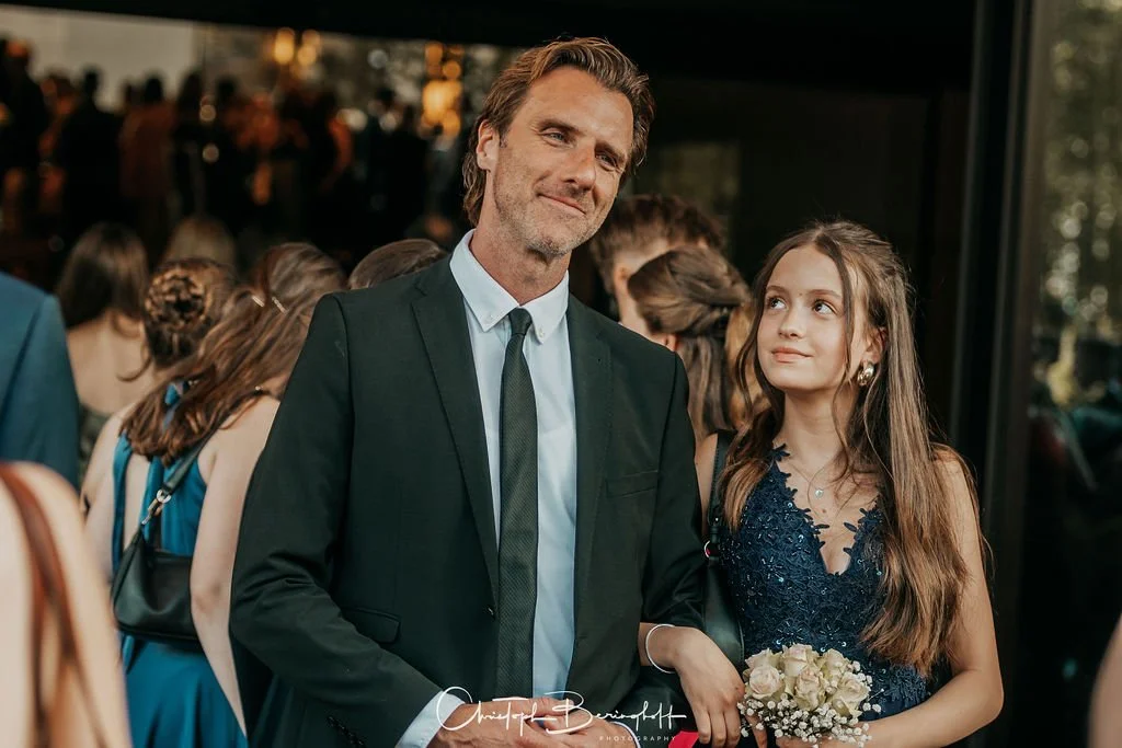 Ein Mann in einem schwarzen Anzug und eine junge Frau in einem blauen Kleid stehen zusammen bei einer formellen Veranstaltung.