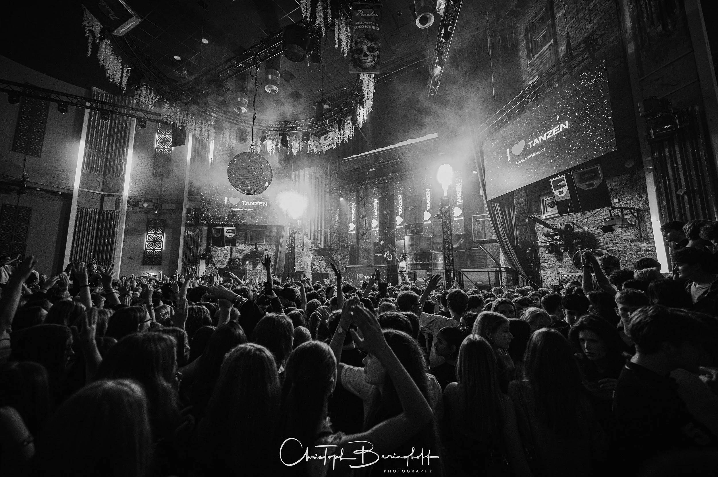 Eine große Menschenmenge feiert in einem dunklen, mit Lichtern beleuchteten Tanzclub. Es gibt eine Bühne mit einem DJ und großen Bildschirmen, die 