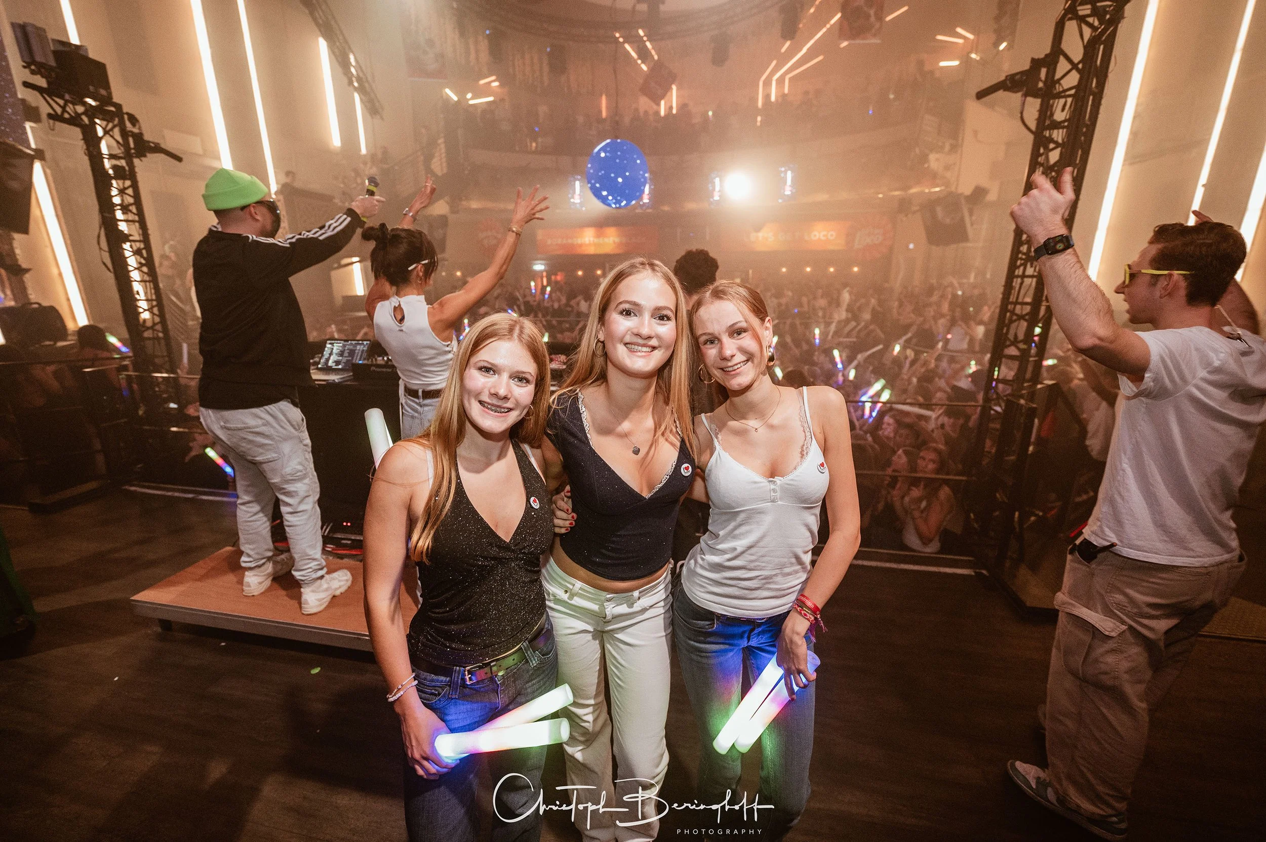 Drei lachende junge Frauen posieren im Mittelpunkt einer Partyszene mit einem DJ und einem tanzenden Publikum. Die Frauen halten Leuchtstäbe. Die Atmosphäre ist lebhaft mit bunten Lichtstrahlen und einer Club-Umgebung.
