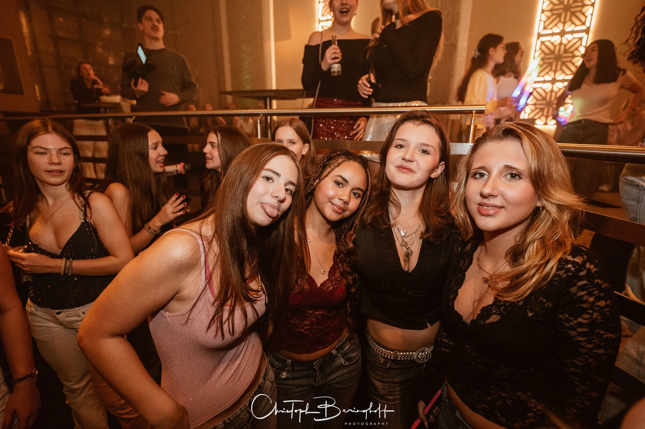 Gruppe junger Frauen beim Feiern in einer Bar oder einem Club, im Hintergrund weitere Gäste und bunte Lichter.