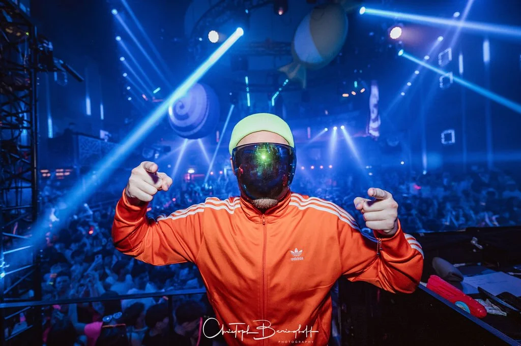 A person in a neon outfit and VR goggles stands in front of a dancing crowd in a club, surrounded by lasers and light effects.