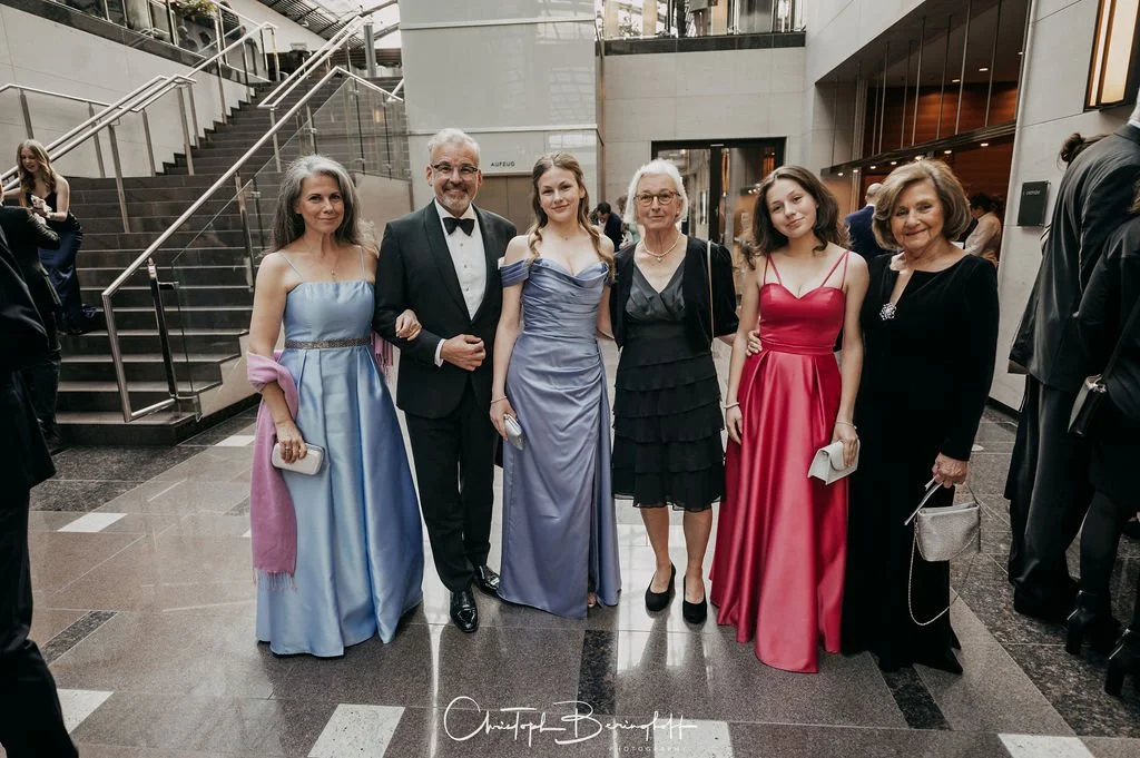 Friedliche Gruppe von sechs Menschen, die formelle Abendkleidung tragen, steht im Eingangsbereich eines modernen Gebäudes mit Treppe im Hintergrund bei einer Veranstaltung.