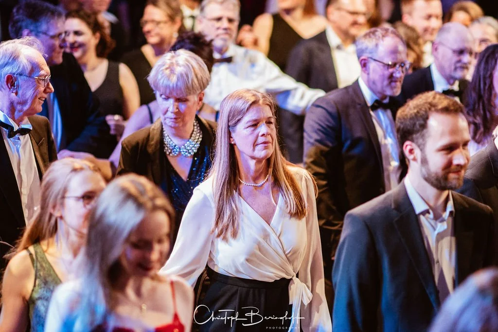 Menschen in formeller Kleidung bei einer Veranstaltung, die meisten tragen Anzüge oder elegante Kleider.