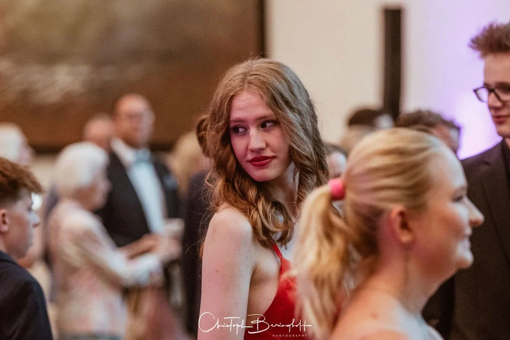 Junge Frau mit lockigem braunem Haar in rotem Kleid, umgeben von anderen Menschen, bei einer formellen Veranstaltung oder Feier.