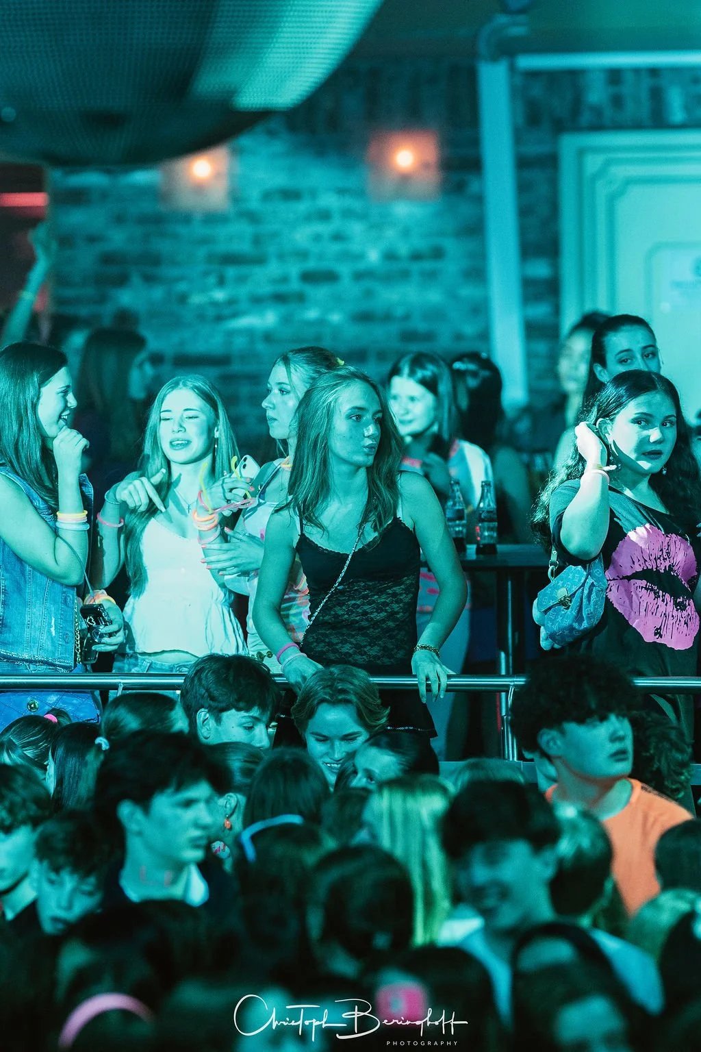 A crowd at a party or concert, mostly teenagers, with bright and dim lights in a room with a brick wall; some are standing by a railing, while others are sitting in the foreground.