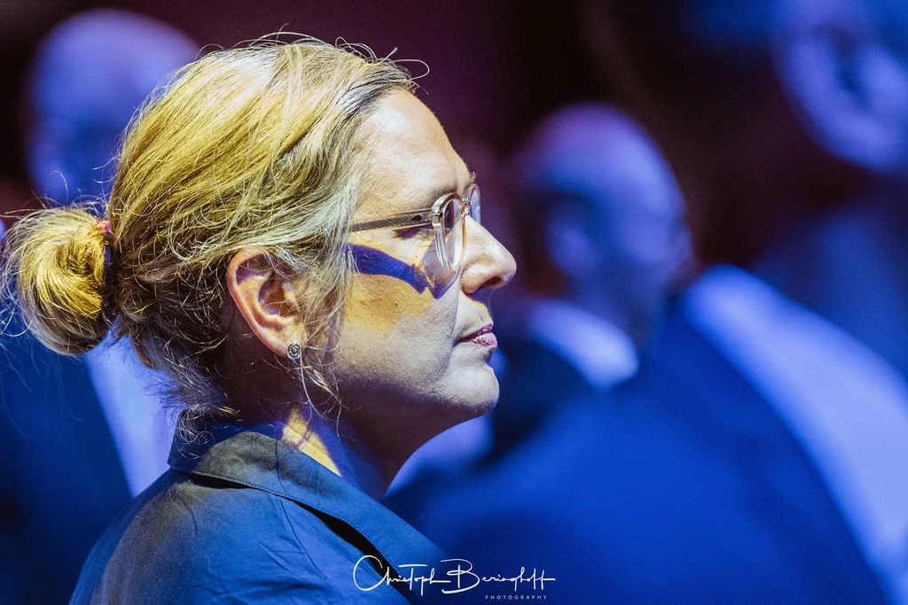 Frau mit grauen Haaren und Brille bei einer Veranstaltung, im Hintergrund weitere Menschen, dunkle ungeblendete Beleuchtung.