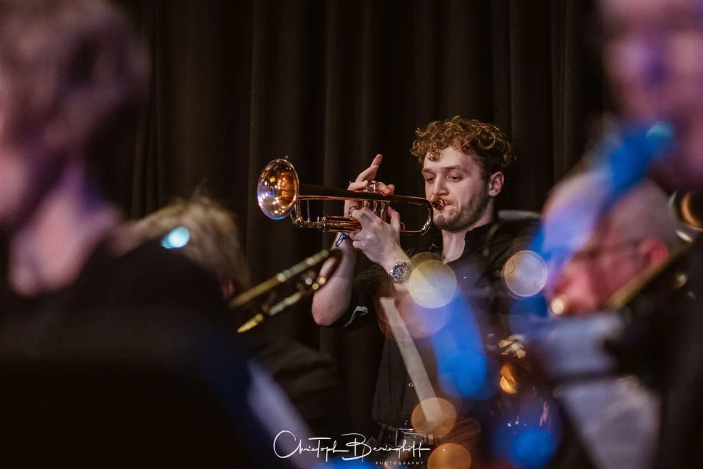 Ein junger Mann spielt Trompete während eines Orchesters auf einer Bühne mit dunklem Vorhang im Hintergrund.