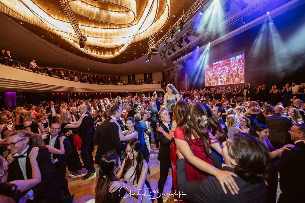 Tanzende Menschen auf einer festlichen Ballveranstaltung mit elegantem Ambiente, Bühnenbeleuchtung und großem Bildschirm im Hintergrund.