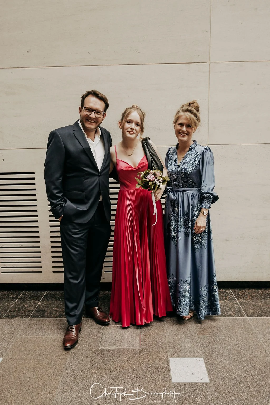 Drei Menschen, zwei Frauen und ein Mann, stehen gemeinsam vor einer Wand. Die Frau in der Mitte trägt ein rotes Kleid und hält einen Blumenstrauß, während die anderen beiden formelle Kleidung tragen.