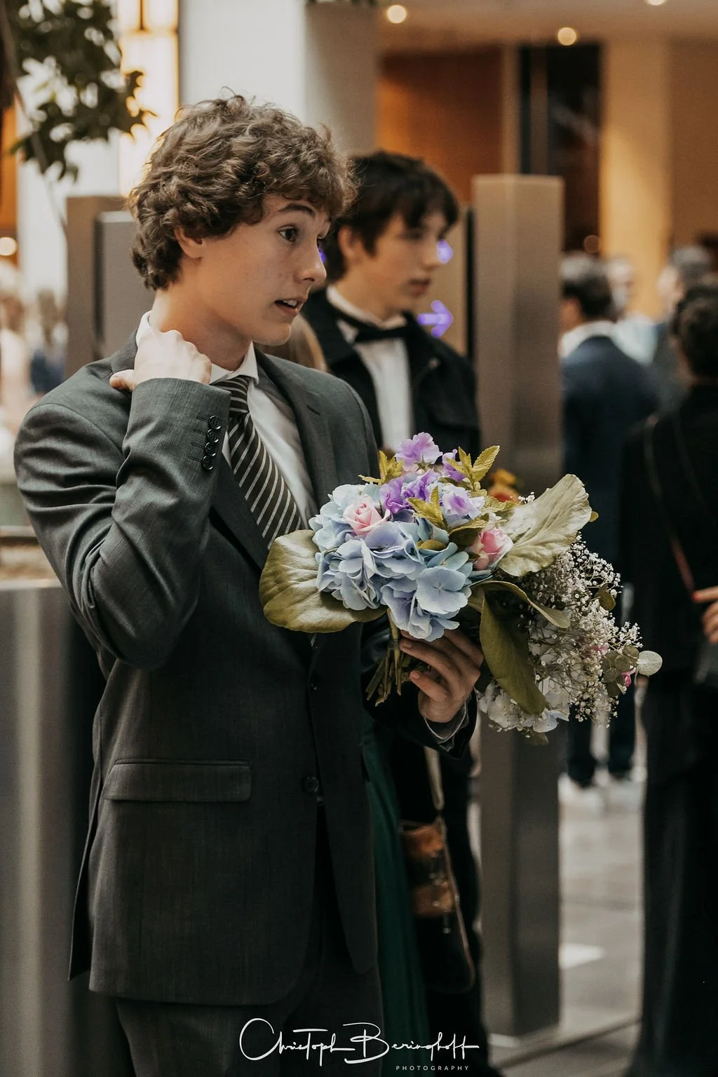 Ein junger Mann in Anzug hält ein Blumenbouquet bei einer formellen Veranstaltung.