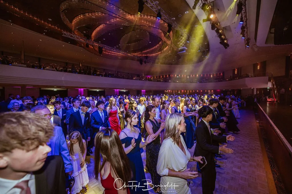 Menschen bei einer formellen Veranstaltung oder Gala im Saal, viele in eleganten Kleidern und Anzügen, mit bunten Lichtern und einer Tanzfläche im Hintergrund.