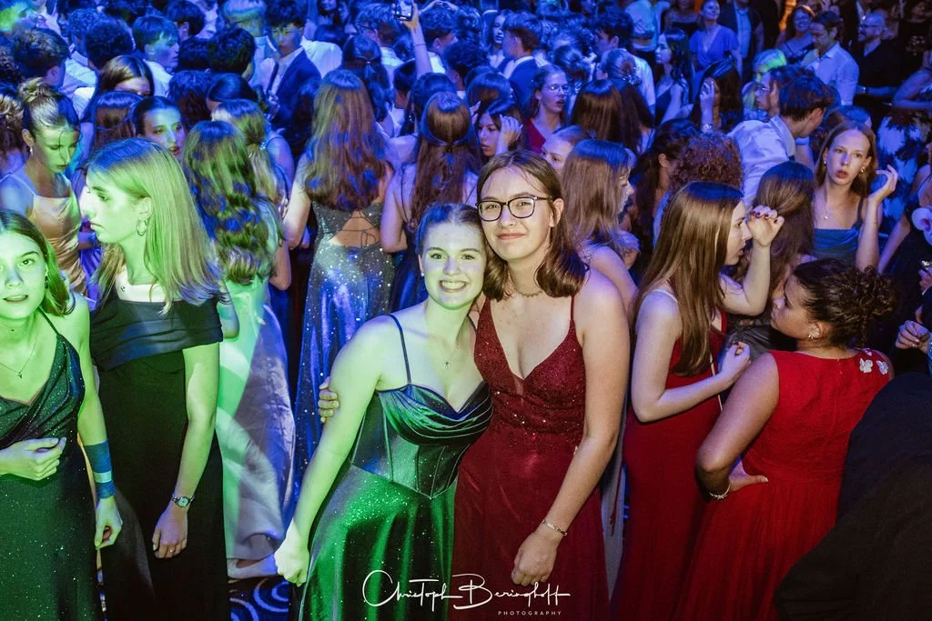 Menschen bei einer festlichen Veranstaltung in eleganter Kleidung, mehrere sitzen und stehen zusammen, im Hintergrund ein Ballsaal mit blauem Licht.