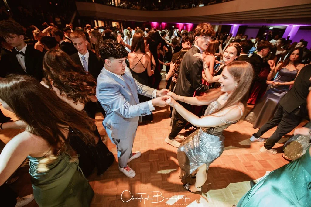 Jugendliche tanzen auf einer Party oder Abschlussball im festlichen, eleganten Outfit, in einem großen Saal mit buntem Licht. Zwei Jugendliche in der Mitte tanzen ineinander.