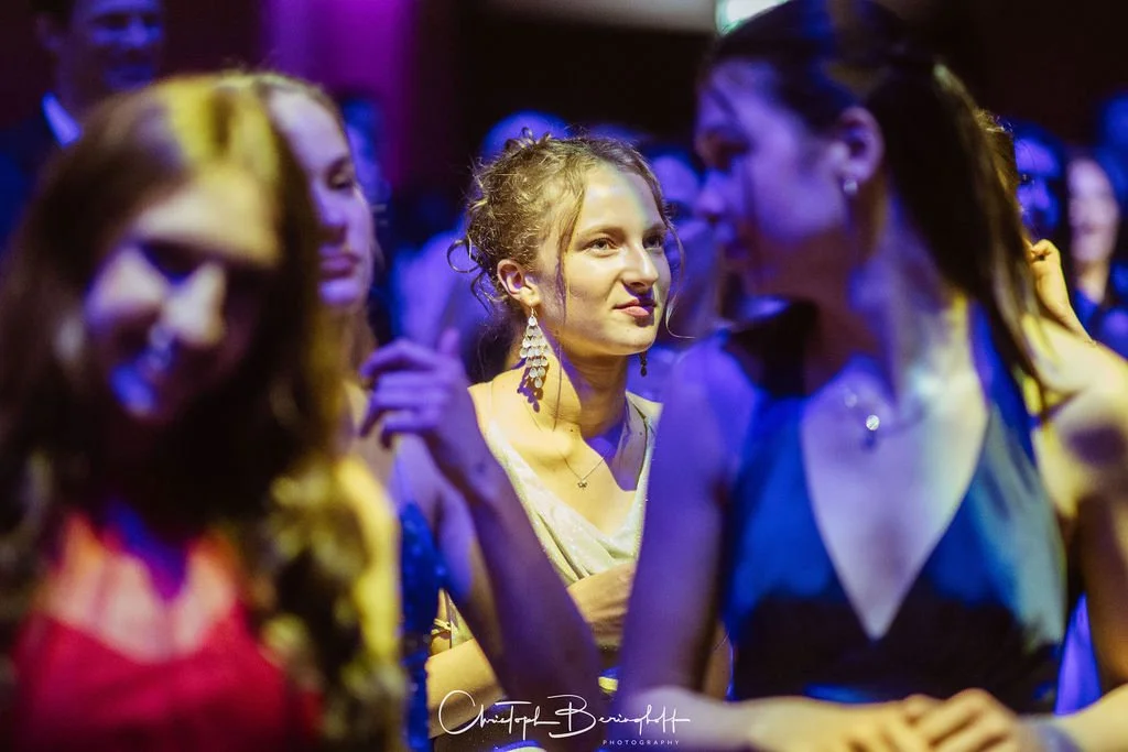 Mehrere Frauen tanzen auf einer Party unter buntem Licht, im Hintergrund dunkle Stimmung.