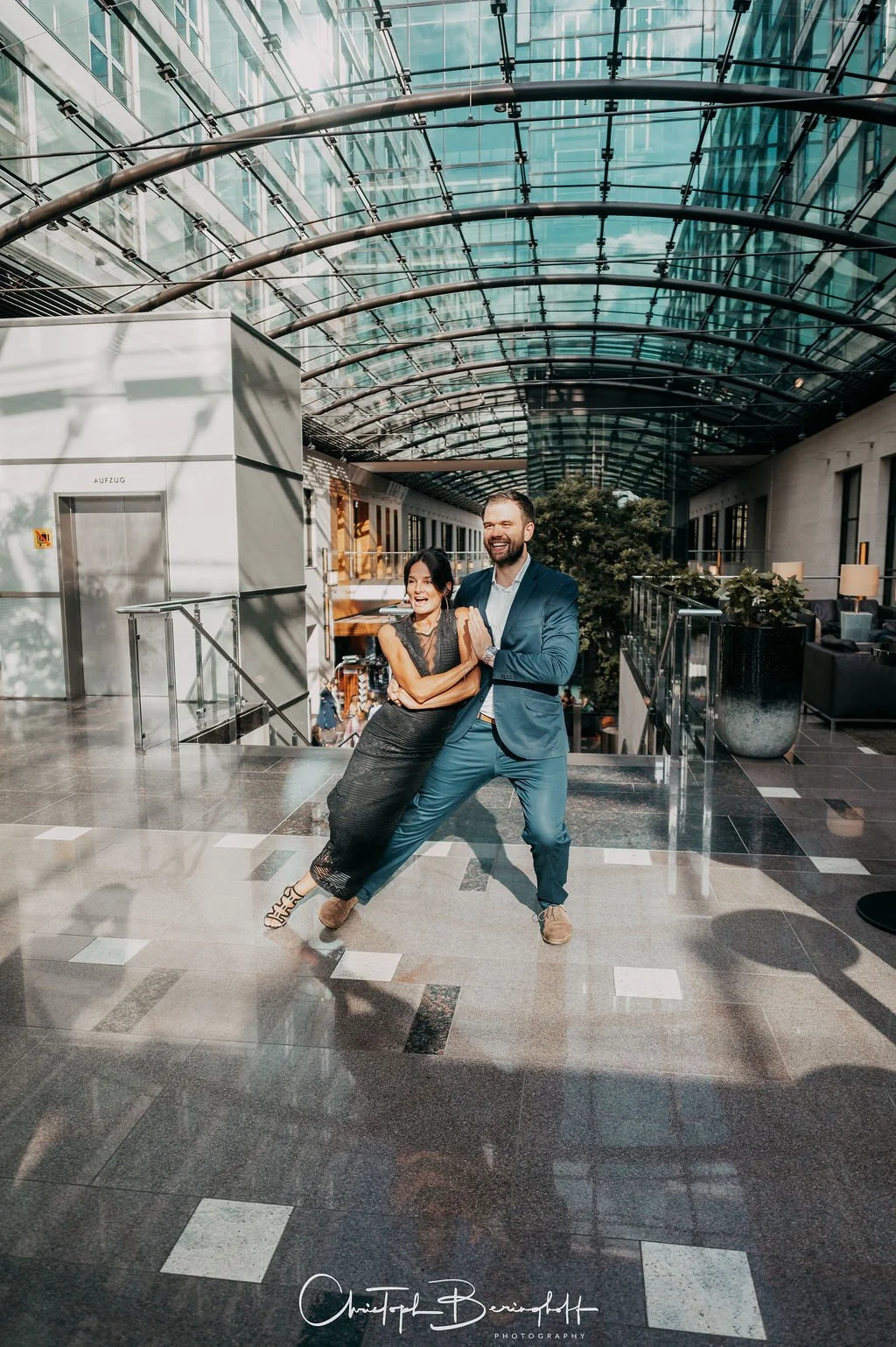 Zwei lachende Menschen, eine Frau und ein Mann, tanzen fröhlich in einem modernen, hellen Atrium mit Glasdach und Steinpflanzen