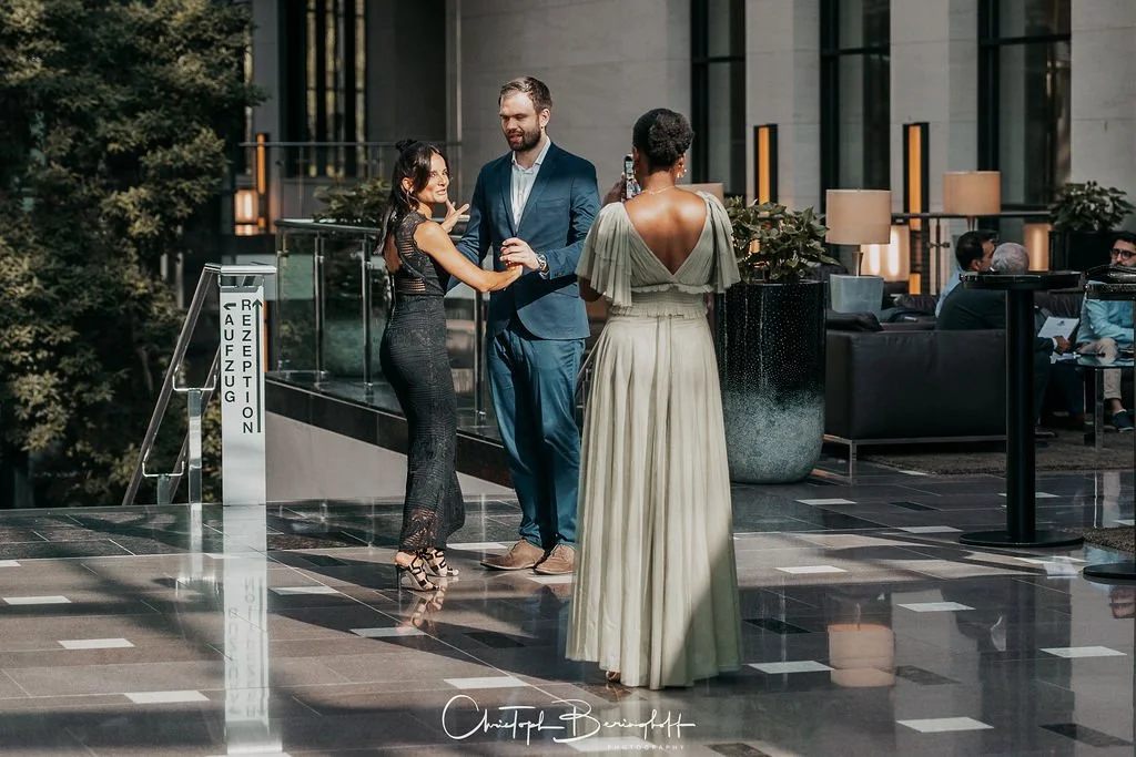 Drei Menschen im formellen Outfit tanzen und lachen in einem eleganten Hotel-Lobby oder Veranstaltungsraum.