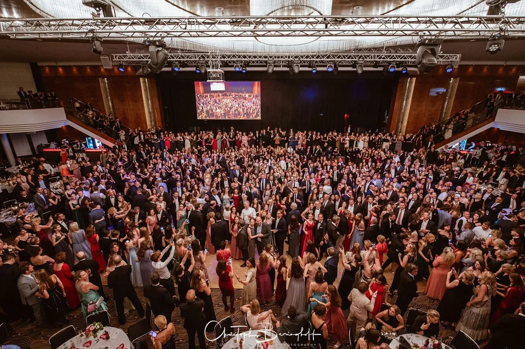Große festliche Veranstaltung in einem Saal mit vielen tanzenden und elegant gekleideten Menschen, einer großen Bühne und einem großen Bildschirm im Hintergrund.